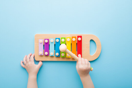 Baby hand holding hammer and playing colorful xylophone on light blue table background. Closeup. Music toy instrument of development for little kids. Point of view shot. Top down view.