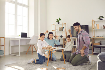 Family weekend entertainment. Mom, dad, daughter and son play game sitting at a table from which a wooden tower falls. Fun board games for family leisure. Stay at home activity for kids.