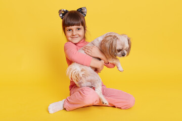 Fototapeta na wymiar Charming little girl and puppy isolated over yellow background, toddler looks directly at camera and embracing her pet with great love, kid wearing hairband with cat's ears.