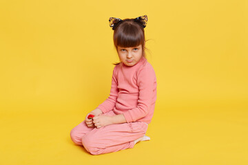 Little dark haired girl looking frowned, sitting on floor against yellow background, toddler wearing casual clothing, offended small kid being upset.