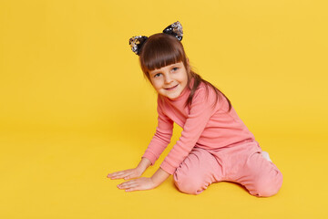 Cute baby girl wearing pink shirt and trousers sitting on floor isolated over yellow background, smiling child in hair band with cat's ears looking directly at camera, touching floor with palms.