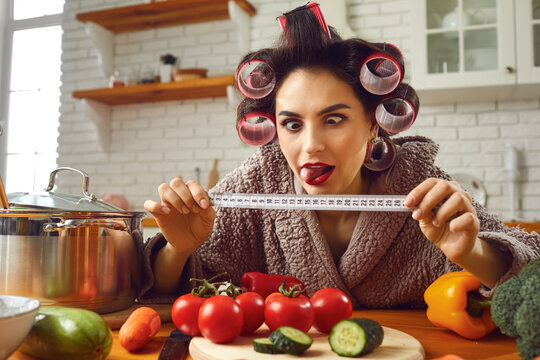 Funny Crazy Young Woman In Hair Rollers Holding Tape Measure In Hands And Looking At The Centimeters She Has To Lose While Cooking Healthy Food In The Kitchen. Weight Loss And Dieting Humorous Concept