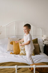 Obraz na płótnie Canvas Beautiful cute four-year-old boy in white clothes smiles and jumps on the bed on the light background of the house