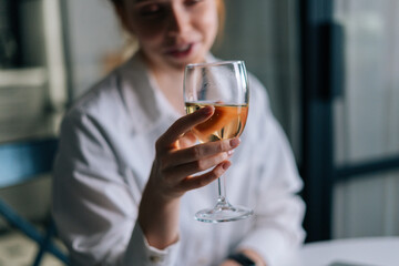 Close-up blurry photo of happy redhead young woman holding glass with champagne near face, looking away. Portrait of pretty female with glass of wine.