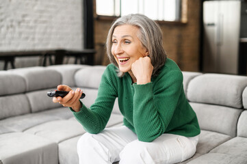 Obraz premium Image of emotional smiling optimistic aged woman grey haired, homely dressed, sitting on comfortable sofa with a TV remote in the hand, watch funny program on TV or excited film, spending time at home