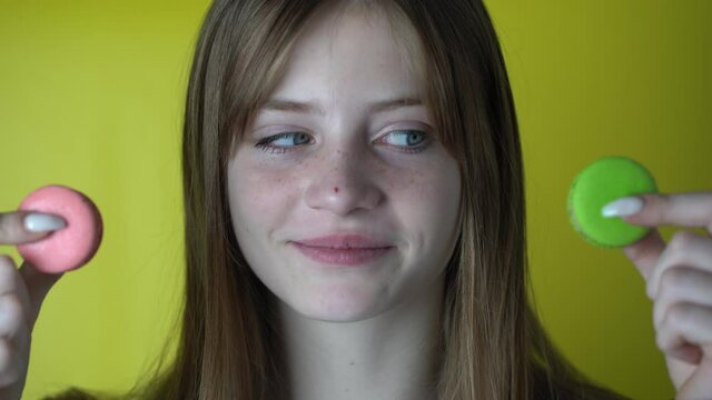Attractive smiling young blond girl is looks at macaroons and does not know which one to choose. Yellow background with copy space. Teenager emotions, indoors, closeup portrait