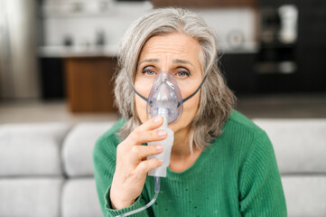 Elderly woman grey-haired, has a lot of wrinkles, feels bad, holding oxygen mask, lacking of...