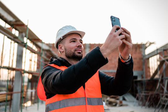Civil engineer or architect with hardhat and orange construction vest on construction site taking photos of the progress with the smartphone camera for the report.
