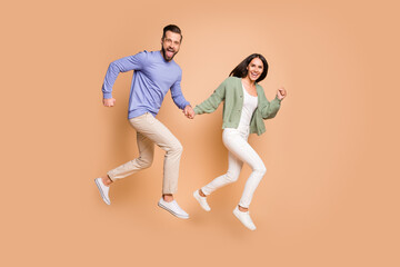 Fototapeta na wymiar Full length body size view of nice cheerful partners couple jumping holding hands running isolated on beige pastel color background