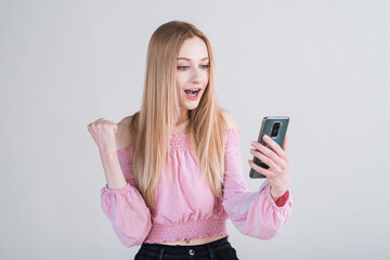 Fototapeta premium Portrait of a blonde girl who holds a smartphone and shows the gesture of the winner in the studio on a white background