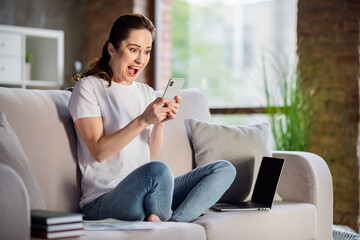 Photo of young shocked amazed surprised businesswoman sit sofa remote working at home hold smartphone see bad news