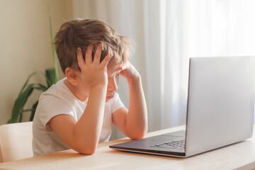 Desperate child sits at desk, looks sadly at computer and holds his head with hands. Online learning problem concept with copy space