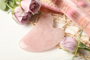 Rose quartz gua sha tool, flowers and cosmetic product on white wooden table, closeup