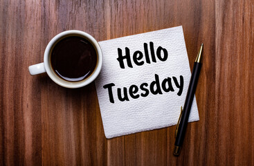 On a wooden table next to a white cup of coffee and a pen is a white paper napkin with the words HELLO TUESDAY
