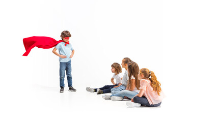 Naklejka premium Rescuer. Child pretending to be a superhero with his friends sitting around him. Kids excited, inspired by their brave friend in red coat isolated on white background. Dreams, emotions concept.