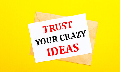 On a yellow background, an envelope and a card with the text TRUST YOUR CRAZY IDEAS. View from above