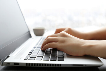 Woman works at laptop on window background. Female hands on keyboard in sunlight
