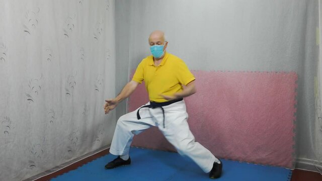 An old man athlete in a yellow T-shirt with a medical mask and with a black belt makes blocks with his hands in a rack