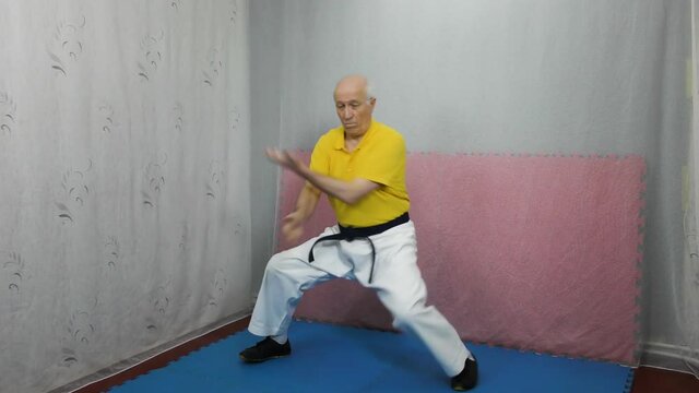 Old man athlete in a yellow T-shirt and with a black belt performs blocks with his hands in a rack