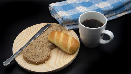 Una taza blanca con cafe, una  tostada de pan con un bollo de leche sobre un plato de madera y una mantel azul sobre fondo negro 