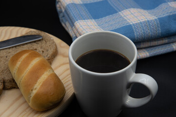 Una taza blanca con cafe, una  tostada de pan con un bollo de leche sobre un plato de madera y una mantel azul sobre fondo negro 