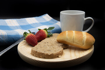 Una taza blanca con cafe, una tostada y un bollo de leche, fresas sobre un plato de madera y un mantel azul, todo sobre fondo negro.