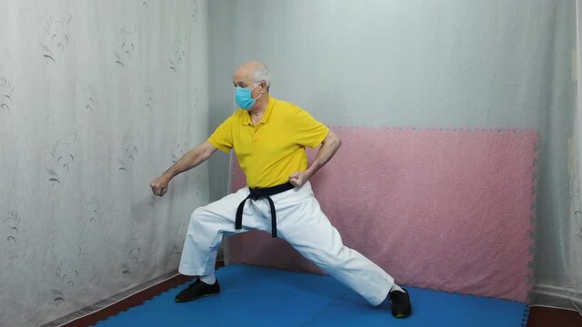 An old man athlete in a yellow T-shirt with a medical mask and with a black belt performs blocks with his hands in a rack