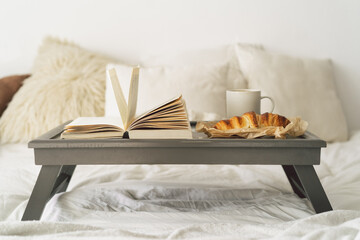 Tray with croissant, cup of coffee and book in white clean bed. Mindful and quiet living.