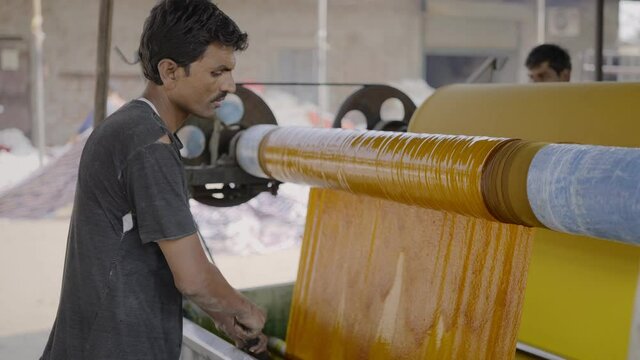 outdoor close shot of a male indian labourer working around a running textile machinery dyeing sheets of clothes, concept of restarting trade of small manufacturing companies amid covid 19 pandemic