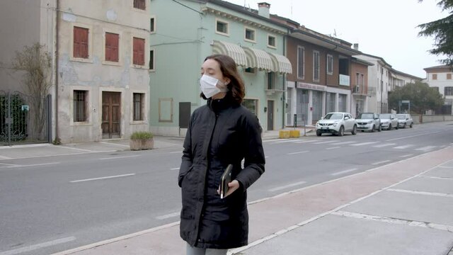 Young girl strolls wearing an anti covid mask. Brunette woman walks the streets of an Italian village. Winter and cloudy sky. Sunday morning in quarantine period.