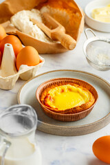 Egg tart, traditional Portuguese dessert, pastel de nata in a gray plate on a marble background . Portuguese puff and ingredients for cooking (flour, butter, eggs, milk). Selective focus