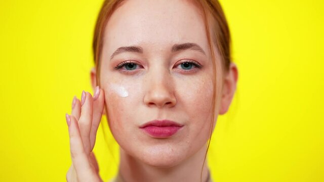 Face of slim freckled redhead young woman with green eyes looking at camera applying skin moisturizer. Close-up portrait of beautiful Caucasian confident lady taking care of beauty.