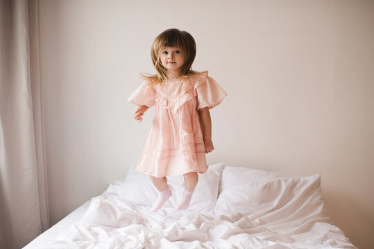 Cute Funny Child Girl 2-3 Year Old Wearing Stylish Pink Dress Jump In Bed In Rom Close Up. Looking At Camera. Childhood. Good Morning. Resting In Hotel Room.