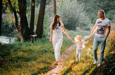Naklejka na ściany i meble A lovely family is walking in the summer forest. Happy parents enjoy the fresh air and beautiful nature, holding hands, talking, smiling and laughing. Healthy lifestyle.