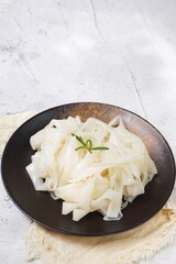 Grainy look Fresh Kwetiau as White Chinese noodles made from Rice in black plate. Popular in South East Asia on texture white background. Selective focus