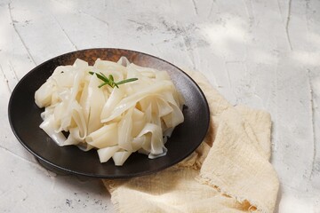 Grainy look Fresh Kwetiau as White Chinese noodles made from Rice in black plate. Popular in South East Asia on texture white background. Selective focus
