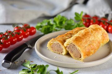 Traditional English pastries - puff pastry rolls with minced meat, on a gray plate, on a gray table with cherry tomatoes and herbs.