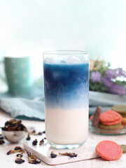 A glass of iced butterfly pea flower milk tea latte  with colorful biscuits and its dried flowers on a table, it's a herbal and caffeine free drink