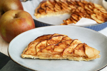 A slice of traditional classic French fine apple tart in a plate made with puff pastry pie, caramelized with brown sugar with belle de boskoop apple in background 