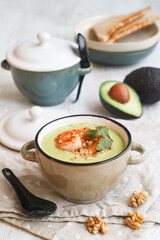 A bowl of healthy chilled avocado soup with grilled shrimps topped with grounded walnuts and coriander leaves in a bowl, with its fruit and toasted bread in background, concept for ketogenic diet food
