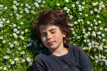Close up portrait of a young boy lying on a glade with daisies and closing eyes with pleasure. Child wearing dark grey t shirt resting on a grass in the park in summer or spring. © Anna