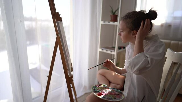 Beautiful portrait of a young girl in a white shirt painting a portrait on canvas. Cute artist sits near the easel and paints with a brush while sitting at home near the window