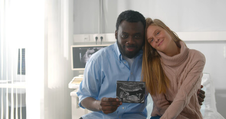 Happy multiethnic couple holding ultrasound scan of their baby sitting in clinic office
