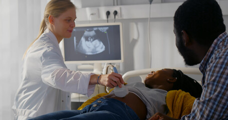 Sonographer scanning and examining african pregnant woman while expecting father looking at monitor