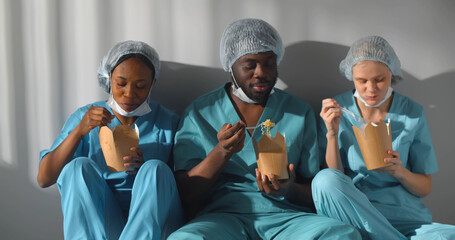 Diverse healthcare workers in scrubs uniform talking and eating takeaway food sitting on floor