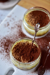 Two jars of cheesecake, sprinkled with cocoa powder, on a white marble board, with a teaspoon.