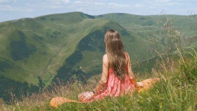 Young woman in red dress sitting on grassy field on a windy day in summer mountains enjoying view of nature.