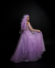 Full length portrait of girl wearing long purple fantasy ball gown with crown and pink hair, standing pose with elegant gestural  movements against a studio background.