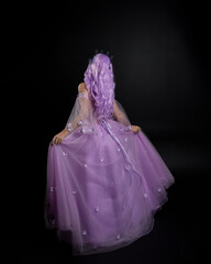 Full length portrait of girl wearing long purple fantasy ball gown with crown and pink hair, standing pose with back to the camera  against a studio background.