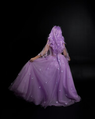 Full length portrait of girl wearing long purple fantasy ball gown with crown and pink hair, standing pose with back to the camera  against a studio background.